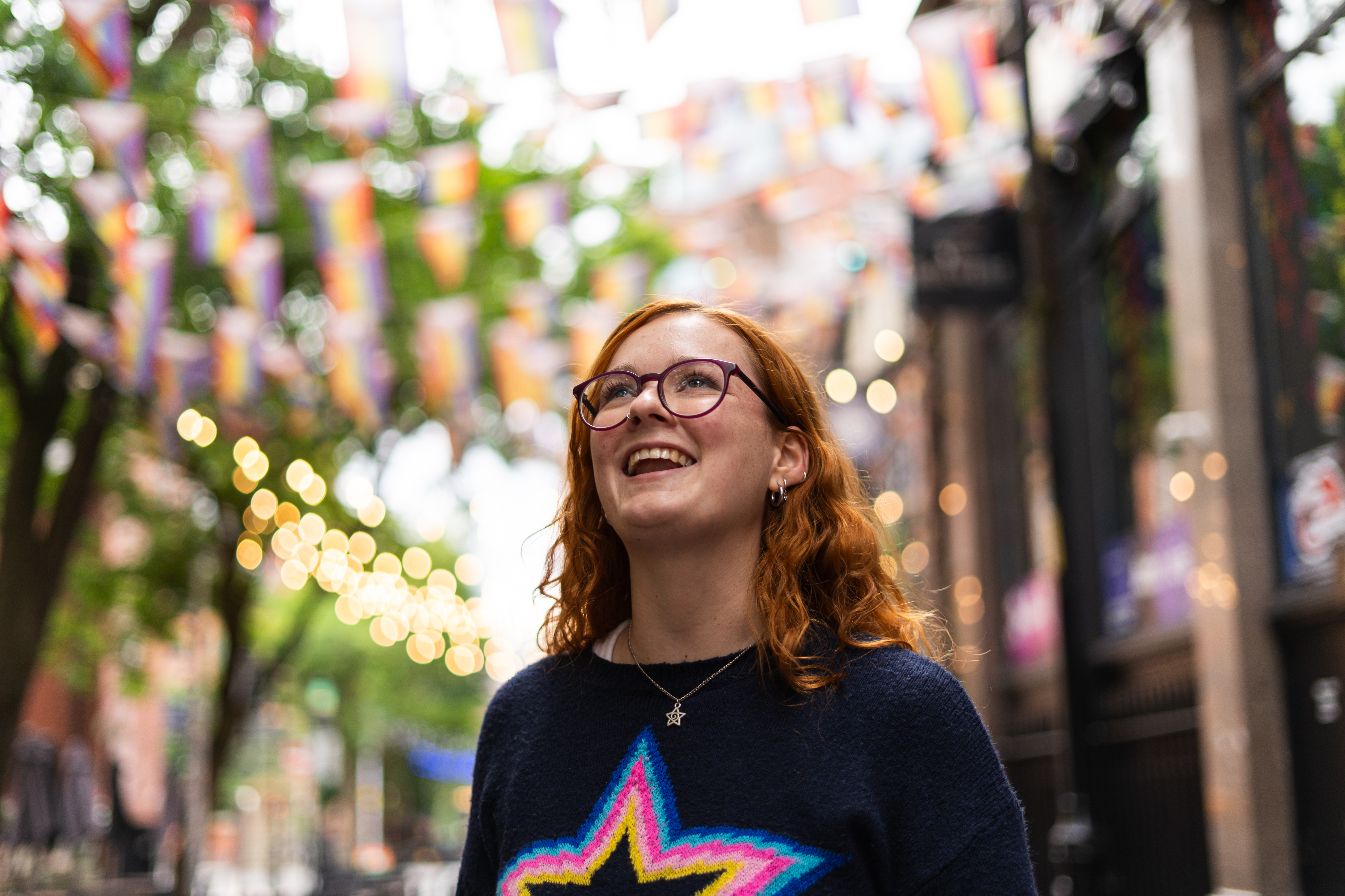 A professional photograph showing a student standing in Manchester's Canal Street, with fairy lights and rainbow flags out-of-focus in the background.
